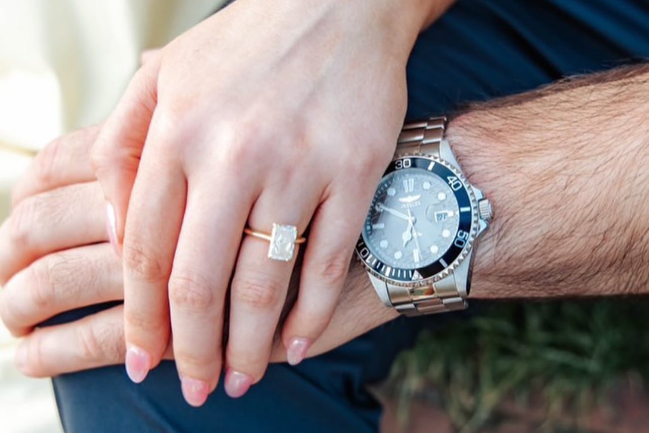 A close-up of a couple holding hands, her wearing a stunning solitaire diamond ring, and him a luxury watch.