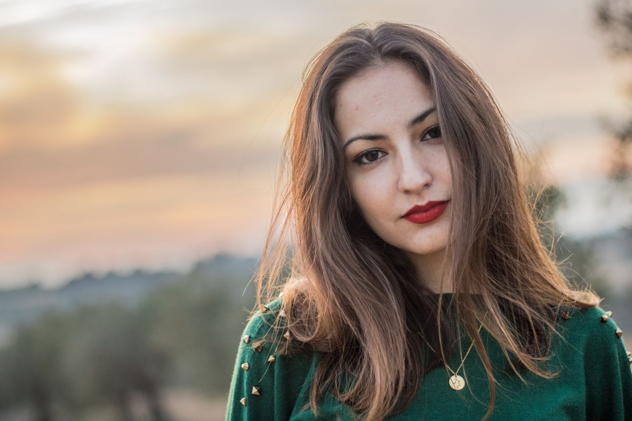A woman in a green top shows off a gold circle necklace while outside at sunset.